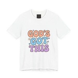 Op de afbeelding: Wit t-shirt met de tekst "GOD'S GOT THIS" in kleurrijke, afgeronde letters. Het ontwerp bevat hartjes, een kruis en een zon. Het t-shirt heeft een ronde hals.