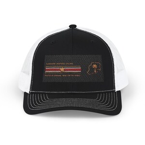 Op de afbeelding: Zwarte en witte truckerpet met een zwart voorpaneel en een witte mesh achterkant. De voorkant heeft een rechthoekige patch met de tekst "SURINAME INSPIRED COLORS" en een ontwerp geïnspireerd op de vlag van Suriname.