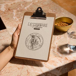 Może przedstawiać: Klip z tekstem "Dinnerware Collection" i ilustracją talerza. Mała złota miska z jedzeniem i szklanka wody znajdują się na marmurowej powierzchni. Klip jest trzymany przez osobę.