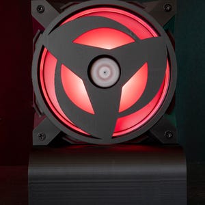 Peut inclure: Un ventilateur de refroidissement pour ordinateur noir doté d'un cadre circulaire avec un motif en forme de shuriken à trois branches. Les pales du ventilateur sont éclairées par une lumière LED rouge vive, le tout monté sur une base rectangulaire noire.