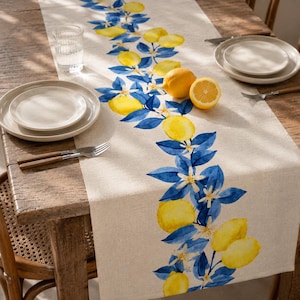 May include: A beige linen table runner with a watercolor design of yellow lemons, blue leaves, and white blossoms. The runner is placed on a wooden table set with plates, silverware, and a glass of water. A whole lemon and a half-cut lemon are also on the table.