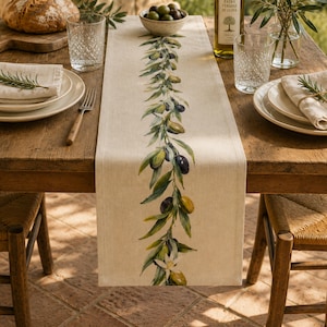 May include: A rectangular beige table runner featuring a central vertical botanical illustration of olive branches with green and dark purple olives, green leaves, and a small white flower, printed on a textured fabric surface.