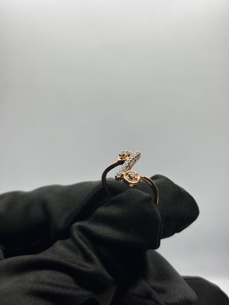 Puede incluir: Un anillo de oro rosa con un dise&ntilde;o de letra "Z" en cursiva, incrustado con peque&ntilde;os diamantes brillantes. El anillo se presenta sobre un fondo blanco liso, resaltando los detalles intrincados de la joya.