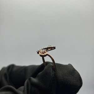 Puede incluir: Un anillo dorado con un dise&ntilde;o &uacute;nico, con peque&ntilde;as piedras brillantes. La banda del anillo se curva con gracia, conduciendo a una secci&oacute;n superior adornada. Las piedras est&aacute;n engastadas en un metal oscuro, creando un contraste con el oro.