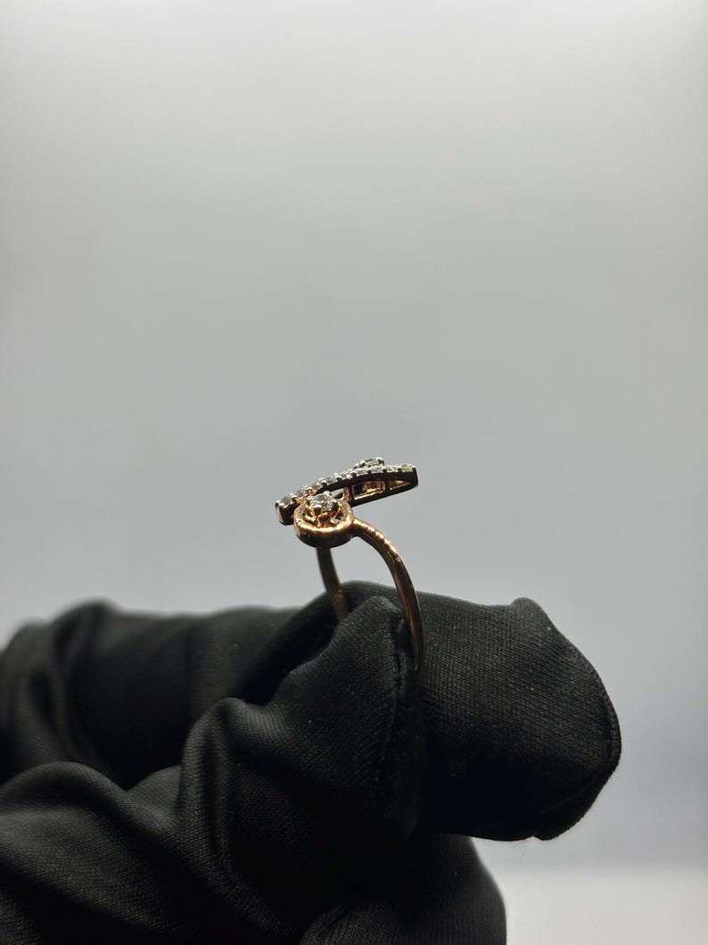 Puede incluir: Un anillo dorado con un dise&ntilde;o &uacute;nico, con peque&ntilde;as piedras brillantes. La banda del anillo se curva con gracia, conduciendo a una secci&oacute;n superior adornada. Las piedras est&aacute;n engastadas en un metal oscuro, creando un contraste con el oro.