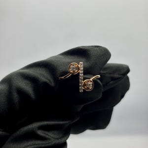 Puede incluir: Un anillo dorado con dos engastes circulares y una barra vertical, todos adornados con peque&ntilde;as piedras preciosas transparentes. El anillo se muestra sobre un fondo negro, resaltando su delicado dise&ntilde;o y brillo. El dise&ntilde;o del anillo es moderno y elegante.