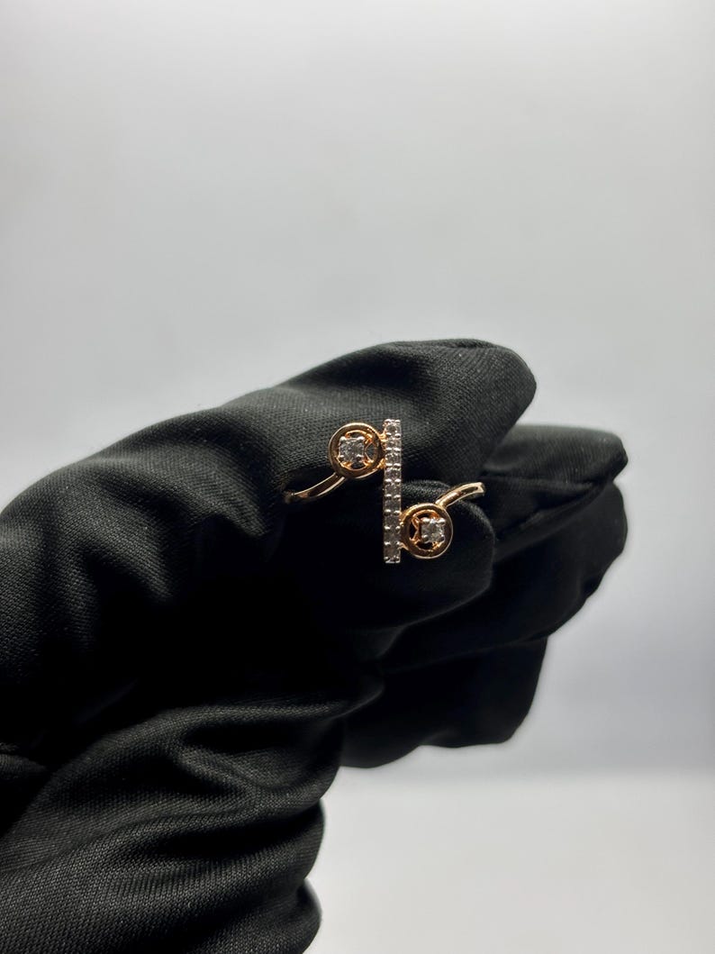 Puede incluir: Un anillo dorado con dos engastes circulares y una barra vertical, todos adornados con peque&ntilde;as piedras preciosas transparentes. El anillo se muestra sobre un fondo negro, resaltando su delicado dise&ntilde;o y brillo. El dise&ntilde;o del anillo es moderno y elegante.