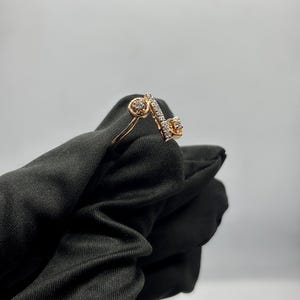 Puede incluir: Un anillo de oro rosa con un dise&ntilde;o &uacute;nico. El anillo presenta dos engastes circulares con peque&ntilde;as piedras y una barra diagonal con una fila de peque&ntilde;as piedras. El anillo est&aacute; sobre un fondo negro.