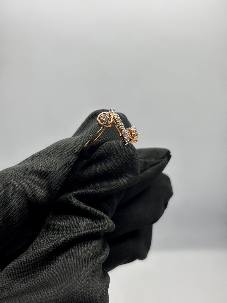 Puede incluir: Un anillo de oro rosa con un dise&ntilde;o &uacute;nico. El anillo presenta dos engastes circulares con peque&ntilde;as piedras y una barra diagonal con una fila de peque&ntilde;as piedras. El anillo est&aacute; sobre un fondo negro.