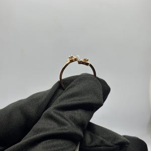 Puede incluir: Un anillo de oro con un dise&ntilde;o abierto &uacute;nico. El anillo presenta peque&ntilde;as piedras preciosas en cada extremo y una l&iacute;nea de piedras m&aacute;s peque&ntilde;as en el centro. El anillo es sostenido por una mano enguantada de negro sobre un fondo blanco.