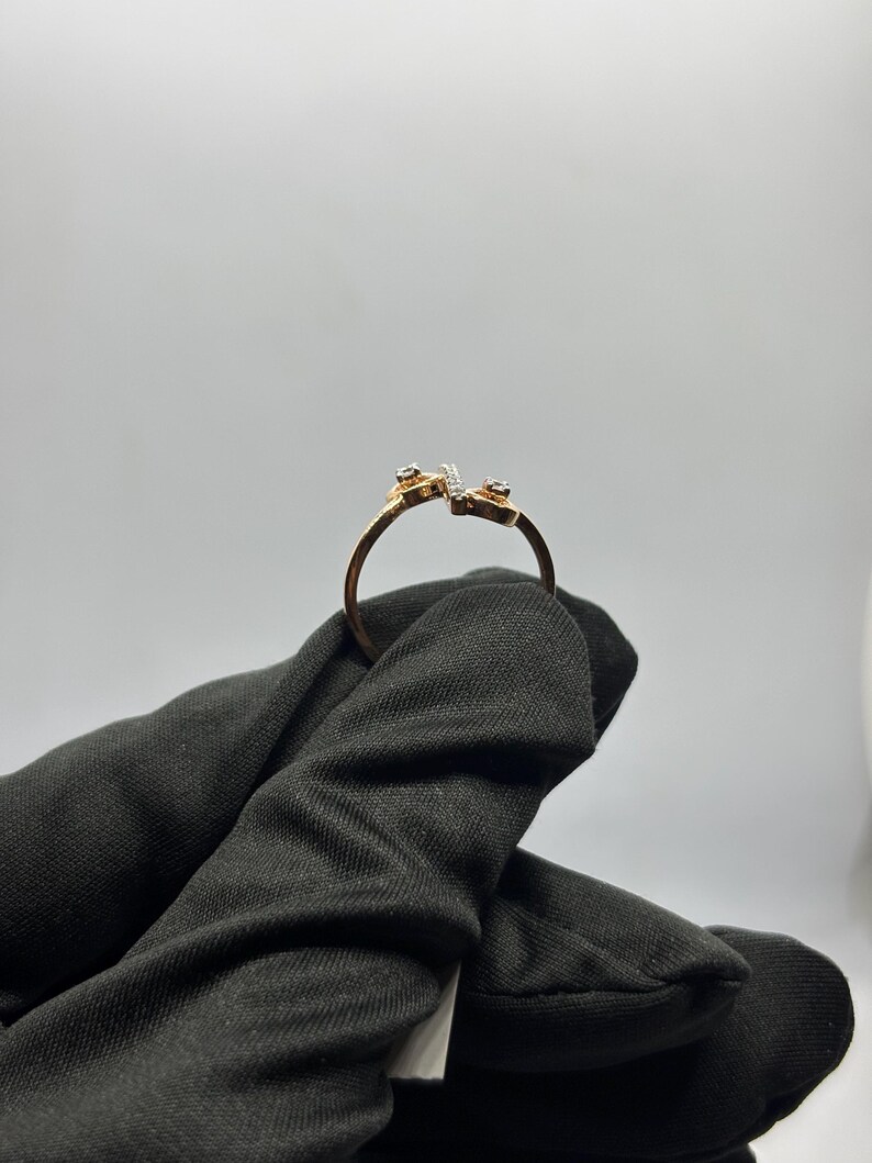 Puede incluir: Un anillo de oro con un dise&ntilde;o abierto &uacute;nico. El anillo presenta peque&ntilde;as piedras preciosas en cada extremo y una l&iacute;nea de piedras m&aacute;s peque&ntilde;as en el centro. El anillo es sostenido por una mano enguantada de negro sobre un fondo blanco.