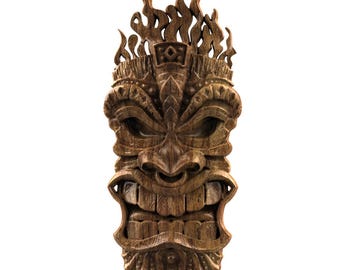 Può includere: Una maschera Tiki in legno dettagliata con un'espressione feroce. La maschera è scolpita in legno marrone scuro, con denti prominenti, sopracciglia aggrottate e un design a forma di fiamma nella parte superiore. È un oggetto decorativo.