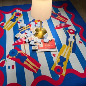 May include: A blue and white striped beach-themed mat with red wavy borders and illustrations of people sunbathing. A set of Mahjong tiles and a game box are on the mat, along with a lamp.