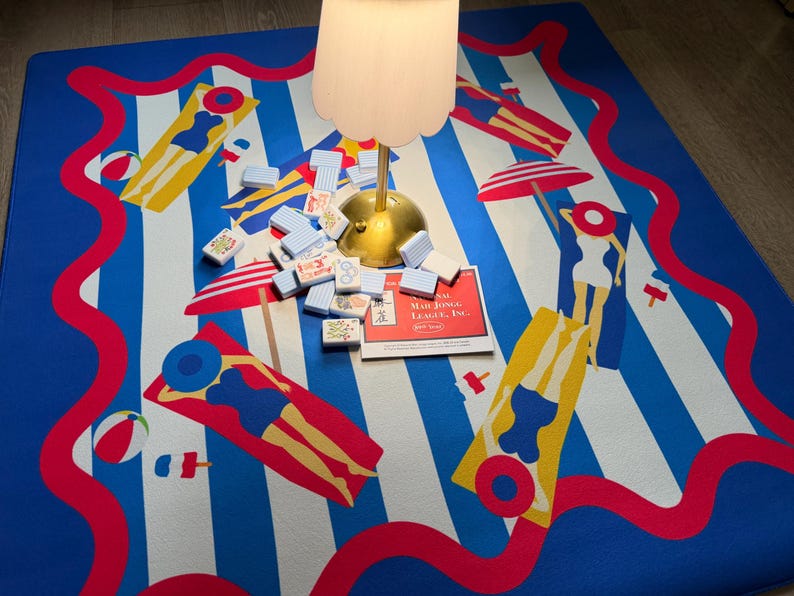 May include: A blue and white striped beach-themed mat with red wavy borders and illustrations of people sunbathing. A set of Mahjong tiles and a game box are on the mat, along with a lamp.
