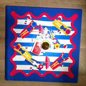 May include: A square mat with a blue border and white and blue stripes. The mat features illustrations of people on floats, beach balls, and other beach-themed items. A small red sign is visible.
