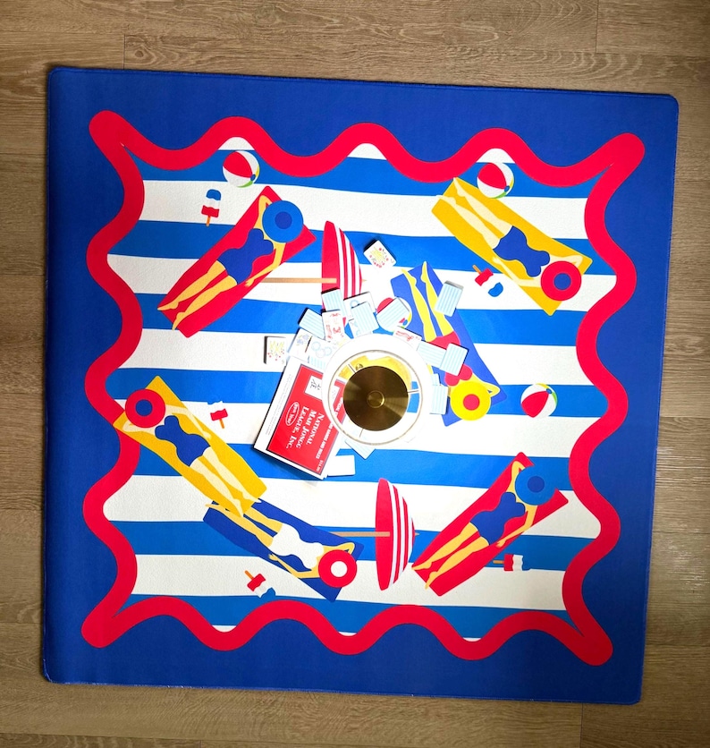 May include: A square mat with a blue border and white and blue stripes. The mat features illustrations of people on floats, beach balls, and other beach-themed items. A small red sign is visible.