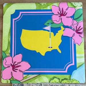 May include: A square decorative mat featuring a blue rectangle with a yellow outline of the United States and a golf flag. Pink flowers and green foliage surround the blue rectangle. The mat has a green and white border.