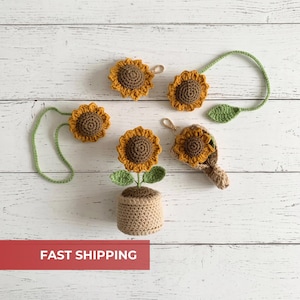 Peut inclure: Diverses décorations de tournesols crochetés, dont des porte-clés et une plante en pot. Les tournesols ont des centres bruns et des pétales jaunes, avec des tiges et des feuilles vertes. Les objets sont disposés sur une surface en bois blanc.