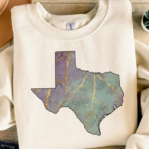 May include: Cream-colored sweatshirt featuring a graphic of the state of Texas. The Texas outline is filled with a marbled design in shades of purple, teal, and gold. The sweatshirt has a crew neck and long sleeves.