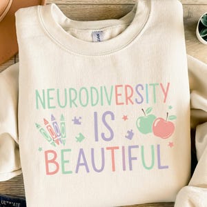 Könnte beinhalten: Cremefarbenes Sweatshirt mit den Worten "NEURODIVERSITY IS BEAUTIFUL" in Pastellfarben. Das Design umfasst Buntstifte, Puzzleteile, Äpfel und Sterne. Das Sweatshirt ist aus weichem Material.