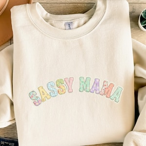 Könnte beinhalten: Cremefarbenes Sweatshirt mit dem Text "SASSY MAMA" in pastellfarbenen Patchwork-Buchstaben. Die Buchstaben sind in Blau-, Rosa-, Grün- und Gelbtönen gehalten. Das Sweatshirt hat einen Rundhalsausschnitt und lange Ärmel.