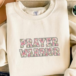 Könnte beinhalten: Cremefarbenes Sweatshirt mit dem Schriftzug "PRAYER WARRIOR" in einem floralen Design. Der Text ist in einem Blockbuchstabenstil mit rosa Rosen und grünen Blättern gehalten. Das Sweatshirt hat einen Rundhalsausschnitt.