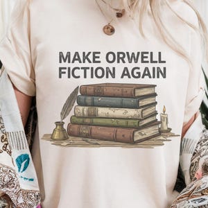 Könnte beinhalten: Beiges T-Shirt mit dem grauen Schriftzug "MAKE ORWELL FICTION AGAIN". Die Grafik zeigt einen Stapel Bücher, Feder und Tintenfass sowie eine brennende Kerze. Das Design hat ein Vintage- und Literaturthema.