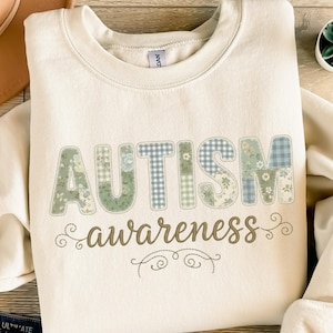 Könnte beinhalten: Cremefarbenes Sweatshirt mit den Worten "AUTISM" und "awareness" im Patchwork-Design. Die Buchstaben sind in Grün- und Blautönen mit Blumen- und Vichy-Mustern gehalten. Das Wort "awareness" ist in Schreibschrift gehalten.