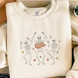Könnte beinhalten: Cremefarbenes Sweatshirt mit einer Grafik von drei tanzenden Skeletten. Das mittlere Skelett trägt den Text "FUCK TRUMP" in Orange. Die Skelette sind von orangefarbenen und gelben Sternen und Blumen umgeben. Das Sweatshirt ist ein lässiger Stil.