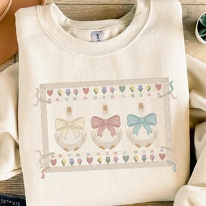 Könnte beinhalten: Cremefarbenes Sweatshirt mit einem verspielten Design von drei Gänsen, die jeweils mit einer großen Schleife in Pastelltönen von Gelb, Rosa und Blau verziert sind. Die Gänse sind von einem dekorativen Rahmen mit Herzen und floralen Motiven umgeben. Ein gemütliches Kleidungsstück.