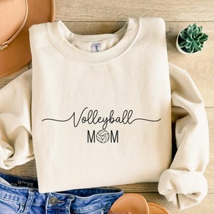 Könnte beinhalten: Cremefarbenes Sweatshirt mit dem schwarzen Schriftzug "Volleyball MOM". Zwischen den Wörtern befindet sich eine Volleyball-Grafik. Das Sweatshirt liegt auf einer Holzoberfläche, im Hintergrund sind eine Jeans und ein brauner Hut zu sehen.