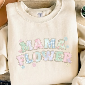Könnte beinhalten: Cremefarbenes Sweatshirt mit dem Schriftzug "MAMA FLOWER" in Pastellfarben. Die Buchstaben sind mit Blumen- und Vichy-Mustern verziert. Kleine Blumengrafiken umgeben den Text und schaffen ein fröhliches Design.