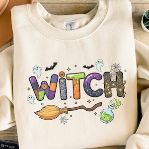 Könnte beinhalten: Cremefarbenes Sweatshirt mit einem farbenfrohen "WITCH"-Aufdruck. Das Design umfasst einen Hexenbesen, Fledermäuse, Geister, ein Spinnennetz und eine Zaubertrankflasche. Die Buchstaben sind in verschiedenen Farben gehalten, darunter Lila, Grün und Orange, mit Patchwork-Effekt auf dem "W".