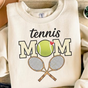 Könnte beinhalten: Cremefarbenes Sweatshirt mit den Worten "tennis MOM" in schwarzen und beigen Blockbuchstaben, einem Tennisball und einem roten Herzen. Zwei gekreuzte Tennisschläger befinden sich darunter. Das Sweatshirt hat einen Rundhalsausschnitt.