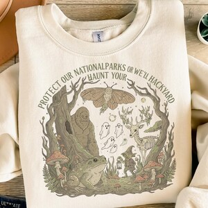 Könnte beinhalten: Cremefarbenes Sweatshirt mit einem Naturmotiv. Das Design zeigt einen Bigfoot, Geister, einen Frosch, Pilze und ein Hirschskelett in einer Waldszene. Der Text um das Bild lautet: "PROTECT OUR NATIONAL PARKS OR WE'LL HAUNT YOUR HACKYARD."