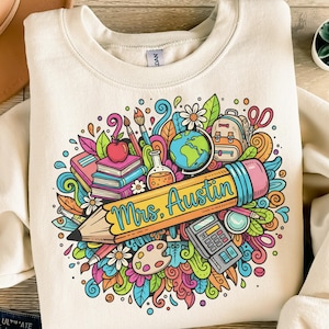 Può includere: Felpa color crema con una grafica colorata a tema insegnante. Il design include una grande matita con la scritta "Mrs. Austin", circondata da materiale scolastico come libri, una mela, un mappamondo e uno zaino.