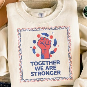 Könnte beinhalten: Cremefarbenes Sweatshirt mit roter, weißer und blauer Grafik. Die Grafik zeigt eine geballte Faust, umgeben von Kreisen und Sternen, innerhalb eines dekorativen Rahmens. Der Text "TOGETHER WE ARE STRONGER" steht darunter.