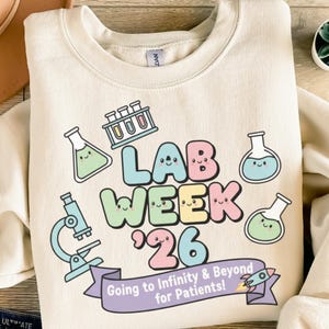 Könnte beinhalten: Cremefarbenes Sweatshirt mit farbenfrohem Grafikdesign. Das Design enthält den Text "LAB WEEK '26" mit lächelnden Laborgeräte-Illustrationen. Ein Banner lautet "Going to Infinity & Beyond for Patients!" mit einer Rakete.