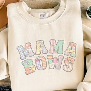 Könnte beinhalten: Cremefarbenes Sweatshirt mit dem Text "MAMA BOWS" in einem Patchwork-Design. Die Buchstaben sind in Pastellfarben mit Schleifenakzenten gehalten, was eine weiche und feminine Ästhetik erzeugt. Das Sweatshirt hat einen Rundhalsausschnitt.