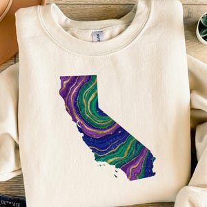 Könnte beinhalten: Cremefarbenes Sweatshirt mit einer farbenfrohen Umrisslinie des Bundesstaates Kalifornien. Das Design innerhalb der Umrisslinie hat einen Marmoreffekt mit Farbtönen von Lila, Blau, Grün und Gold. Das Sweatshirt hat einen Rundhalsausschnitt und lange Ärmel.
