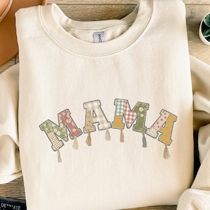 Könnte beinhalten: Cremefarbenes Sweatshirt mit dem Wort "MAMA" in Patchwork-Buchstaben. Jeder Buchstabe hat ein anderes Blumen- oder Karomuster und ist mit einer kleinen Quaste verziert. Das Sweatshirt hat einen Rundhalsausschnitt und lange Ärmel.