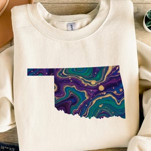 Könnte beinhalten: Cremefarbenes Sweatshirt mit farbenfroher Oklahoma-Umrisszeichnung. Die Grafik des Bundesstaates ist mit einem Wirbelmuster aus Türkis, Lila und Gold gefüllt. Das Sweatshirt hat einen Rundhalsausschnitt und lange Ärmel.