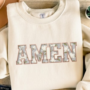 Könnte beinhalten: Cremefarbenes Sweatshirt mit dem Wort "AMEN" in großen Blockbuchstaben. Die Buchstaben haben ein Blumenmuster mit hellgrauem Hintergrund und roségoldener Umrandung. Das Sweatshirt hat einen Rundhalsausschnitt.