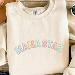 May include: Cream-colored sweatshirt with the text "MAMA STAR" in pastel colors. The letters have a textured, patterned design. The sweatshirt has a crew neck and long sleeves, perfect for casual wear.