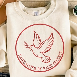 Könnte beinhalten: Cremefarbenes Sweatshirt mit einer roten Taube, die einen Olivenzweig in einem roten Kreis trägt. Die Worte "RADICALIZED BY BASIC DECENCY" sind um den Kreis herum aufgedruckt. Das Sweatshirt hat einen Rundhalsausschnitt und lange Ärmel.