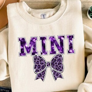 Könnte beinhalten: Cremefarbenes Sweatshirt mit dem Wort "MINI" in großen, lila, edelsteinartigen Buchstaben, die mit silbernem Glitzer umrandet sind. Eine lila Schleife mit Leopardenmuster und Strassakzenten befindet sich darunter. Das Sweatshirt hat einen Rundhalsausschnitt.