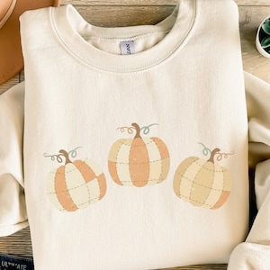Könnte beinhalten: Cremefarbenes Sweatshirt mit drei Kürbis-Illustrationen in Orange- und Cremetönen. Die Kürbisse haben gestickte Details und grüne Ranken, ideal für den Herbst. Ein gemütliches und stilvolles Oberteil zum Thema Herbst.