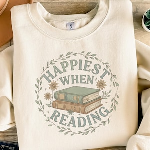 May include: A cream-coloured sweatshirt with a round neckline. The front displays a circular design with the text "Happiest When Reading" in light green. Two stacked books are in the centre, surrounded by floral and leaf accents.