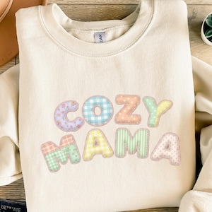 Könnte beinhalten: Cremefarbenes Sweatshirt mit den Worten "COZY MAMA" in bunten Buchstaben im Patchwork-Stil. Die Buchstaben haben Blumen-, Vichy- und Tupfenmuster. Das Sweatshirt hat einen Rundhalsausschnitt.