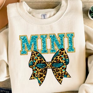 Könnte beinhalten: Cremefarbenes Sweatshirt mit dem Wort "MINI" in türkisfarbenen Glitzerbuchstaben. Eine große Schleife mit Leopardenmuster und türkisfarbenen Akzenten ist unter dem Text zentriert. Die Schleife hat Strassverzierungen.